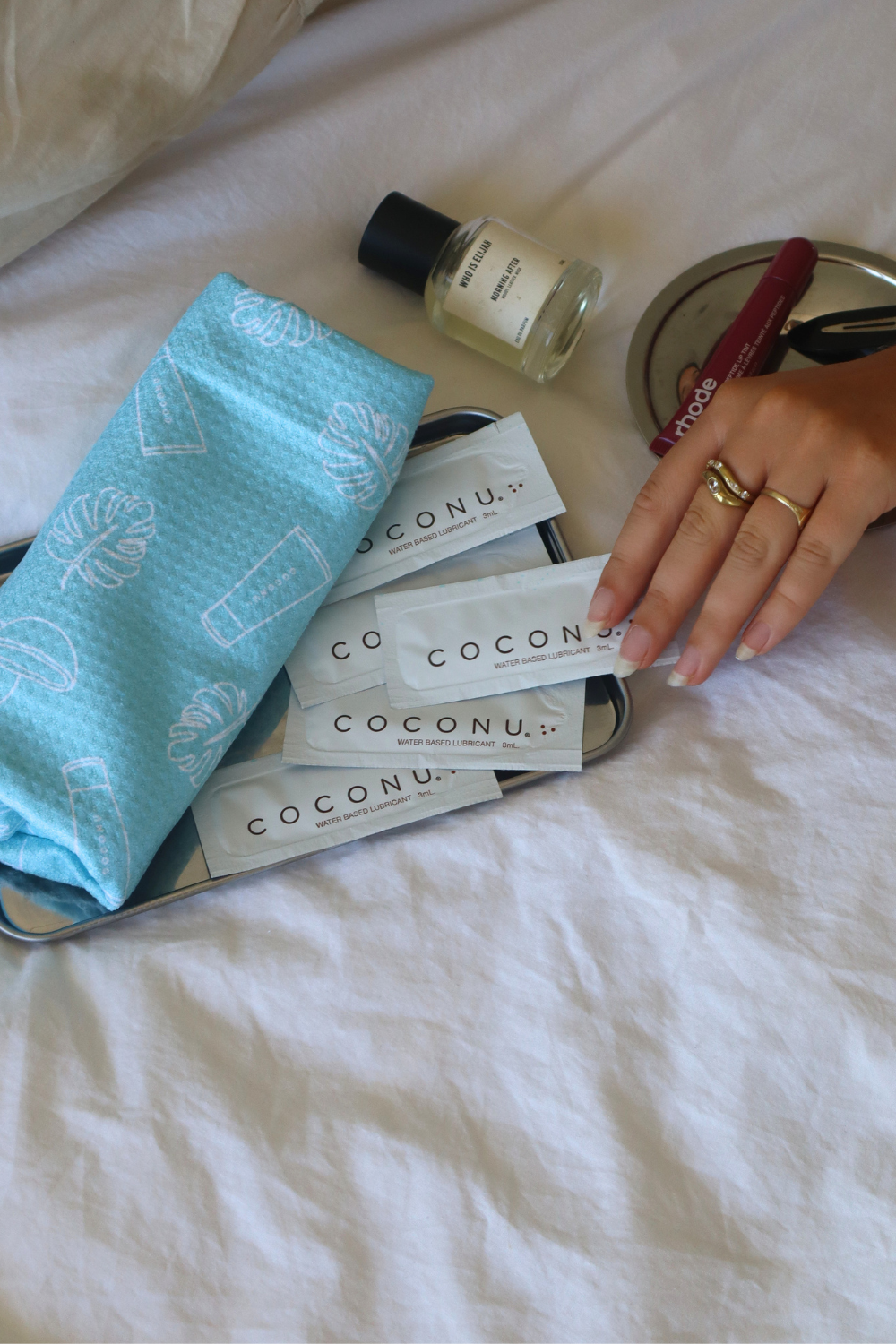 Coconu Play Towel and sachets on white bedding with a perfume bottle